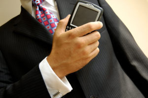 Man in a dark suit and tie, shown from the chest down, holding a BlackBerry phone.
